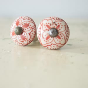 May include: Two ceramic cabinet knobs with a white base and a red floral pattern. Each knob has a silver-colored metal center. The knobs are round and sit on a light-colored surface.