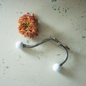 May include: An antique metal wall hook with two white ball ends. The hook has a curved, twisted design and is mounted on a distressed, off-white surface. A dried, orange-toned flower is also present.