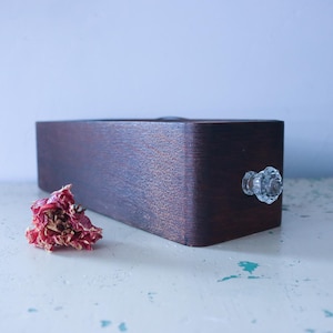 May include: A dark brown wooden box with a clear crystal knob on the right side. A dried red flower sits to the left of the box. The box is rectangular with rounded corners, and sits on a white surface.