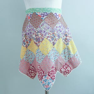 May include: A colorful patchwork skirt with a scalloped hem. The skirt features a variety of patterned fabrics, including polka dots, floral prints, and geometric shapes in shades of yellow, pink, purple, and brown. The waistband is a light green color.