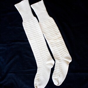 May include: A pair of white knee-high socks made of a thick knit fabric. The socks have a ribbed texture and are laid flat on a black surface.