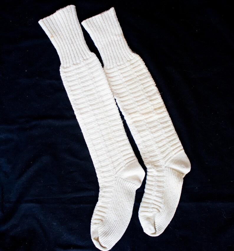 May include: A pair of white knee-high socks made of a thick knit fabric. The socks have a ribbed texture and are laid flat on a black surface.