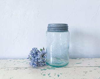 May include: A vintage clear glass jar with a gray metal lid, embossed with text. A small cluster of blue flowers sits beside the jar. The jar is on a distressed white wooden surface against a white wall.
