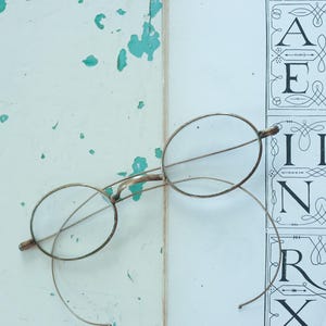 May include: Antique-style oval eyeglasses with thin, metallic gold-toned wire frames and clear lenses. The spectacles feature long, curved temple arms and are positioned over a white surface printed with a black alphabet chart in a serif font.