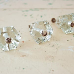May include: Three clear glass cabinet knobs with a hexagonal design and a brown center. The knobs are arranged on a light-colored surface with green paint splatters, suggesting a vintage or antique aesthetic. Ideal for furniture restoration.
