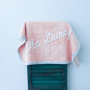 May include: A soft, light pink rectangular towel with a textured surface and a light blue border. The towel features the white embroidered text "The Duncys" in a cursive script across the center, draped over a dark green slatted wooden surface.