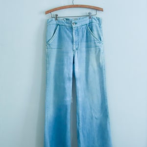 May include: Light blue denim pants with a wide leg design, hanging on a wooden hanger. The pants feature front pockets and a button closure. The denim has a faded appearance.
