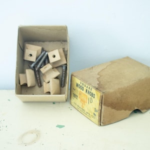 May include: An open cardboard box filled with square wooden knobs and screws sits next to a closed box. The closed box has a yellow label that reads "SQUARE WOOD KNOBS". The knobs are light brown.