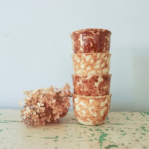 May include: A vertical stack of four small ceramic bowls featuring a mottled, marbled pattern in shades of reddish-brown and cream. Beside the stack of bowls sits a cluster of dried, light brown hydrangea flowers with delicate, papery petals.