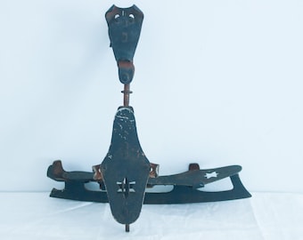 May include: A pair of vintage black metal ice skates with a star design on the blade. The skates have a metal frame and a black leather strap.