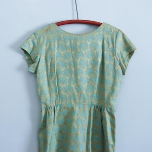 May include: Vintage short-sleeved dress in a light teal color with a gold floral pattern. The dress is hanging on a wooden hanger against a light blue wall. The dress has a fitted waist and a round neckline.