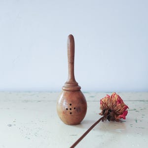 May include: A vintage wooden tea infuser with a long handle and a bulbous body featuring four small holes. A dried red and brown rose rests beside the infuser on a distressed white surface. The tea infuser is a kitchen utensil.