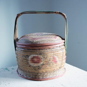 May include: A round, woven basket with a handle and lid. The basket is tan with a floral design in pink and red. The handle is made of a darker material, and the lid has a woven pattern. The basket is sitting on a white surface.