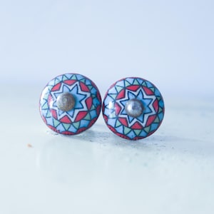 May include: Two round cabinet knobs with a colorful geometric design. Each knob features a central star shape in light blue, surrounded by red, green, and blue patterns. The knobs have a silver-colored metal screw.