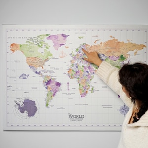 May include: A large world map with a white border, featuring a colorful design with countries in shades of green, purple, and orange. The map includes the text "The World Travel Adventures". A person's hand is pointing at a location on the map.