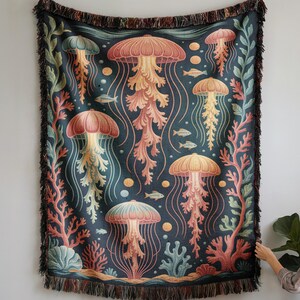 Könnte beinhalten: Eine gewebte Decke mit dunkelblaugrünem Hintergrund, die ein Muster aus Quallen in Korallen-, Pfirsich- und Gelbtönen zeigt. Die Decke mit Fransen wird an einer Wand über einem weißen Sofa präsentiert und verleiht der Einrichtung einen maritimen Touch.