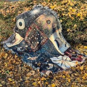 A woven blanket with celestial patterns in dark blue, gold, and cream, featuring suns, moons, and stars, is spread on a bed of fallen autumn leaves.