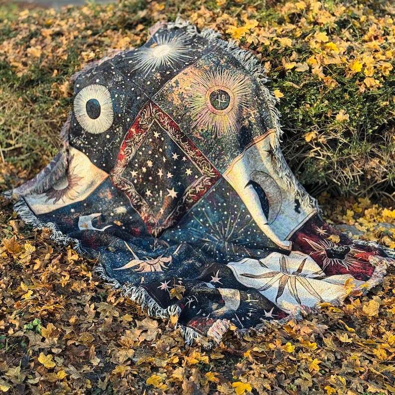 A woven blanket with celestial patterns in dark blue, gold, and cream, featuring suns, moons, and stars, is spread on a bed of fallen autumn leaves.