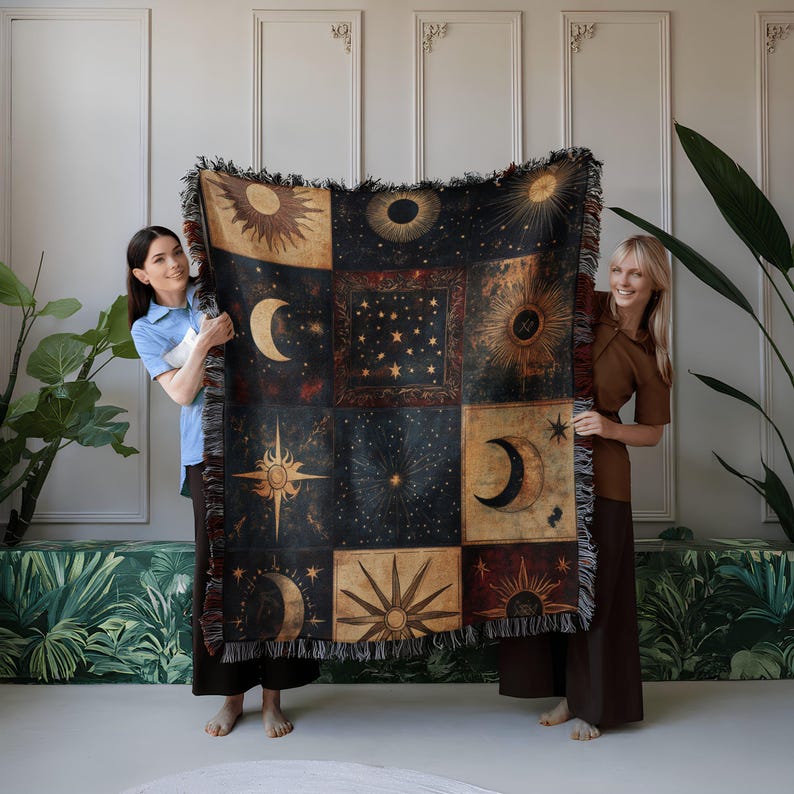 Two women hold up a woven blanket with celestial patterns in dark blue, gold, and cream, featuring suns, moons, and stars, against a white wall.