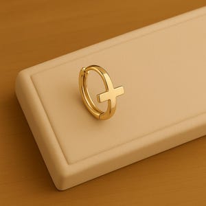 Peut inclure: Boucle d'oreille dorée avec un motif de croix. La boucle d'oreille est en métal brillant et réfléchissant, posée sur un fond neutre. La croix est centrée sur la boucle d'oreille.