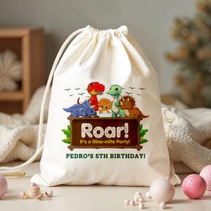 May include: A white drawstring bag featuring a colorful dinosaur graphic. The design includes the word "Roar!" and the text "It's a Dino-mite Party!" and "PEDRO'S 5TH BIRTHDAY!"