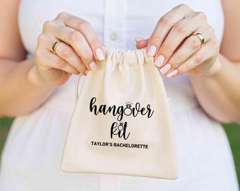 May include: A white cotton drawstring bag with the text "hangover kit" and a ring icon. The text "Taylor's Bachelorette" is printed below the main text.