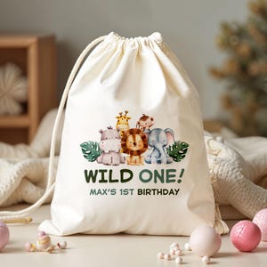 May include: Cream drawstring bag with a watercolour illustration of a lion, elephant, giraffe, monkey, and hippo. Text reads "WILD ONE! MAX'S 1ST BIRTHDAY". The bag is made of a soft, natural fabric.