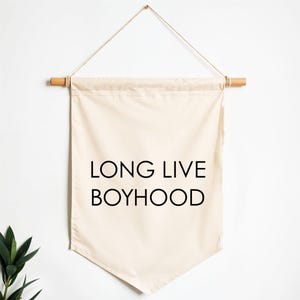 Op de afbeelding: Een beige canvas wandkleed met de tekst "LONG LIVE BOYHOOD" in zwart. De banner is opgehangen aan een houten deuvel met een natuurlijke touwhanger. De banner heeft een puntige onderkant.