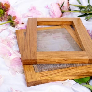 Può includere: Due telai in legno per la fabbricazione della carta con schermi a maglia fine sono impilati su una superficie bianca, circondati da petali e fiori di peonie rosa. I telai sono in legno marrone chiaro e di forma rettangolare.