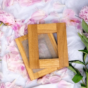 Op de afbeelding: Twee houten frames met een gaasscherm, liggend op een wit oppervlak bezaaid met roze pioenrozenblaadjes. De frames zijn gemaakt van lichtgekleurd hout, met de natuurlijke nerf zichtbaar. De scène is zacht en bloemig.