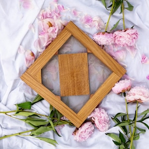 Pode incluir: Uma moldura de madeira com um centro de malha em forma de losango e uma inserção retangular de madeira. Peónias cor-de-rosa e pétalas estão sobre um tecido branco. A madeira é de cor castanho claro.