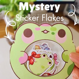 May include: A green frog-shaped container holding a collection of sticker flakes. The container has a light green body with pink accents and brown eyes. The text "Mystery Sticker Flakes" is displayed at the top.