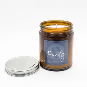 May include: A brown glass candle jar with a silver lid. The candle is white and has a single wick. The label on the jar is blue and says "Purify Clean Burning Soy Candle Hand Poured 6 oz."