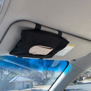 Op de afbeelding: Een zwarte stoffen tissuehouder met een wit tissue erin, bevestigd aan de zonneklep van een auto.