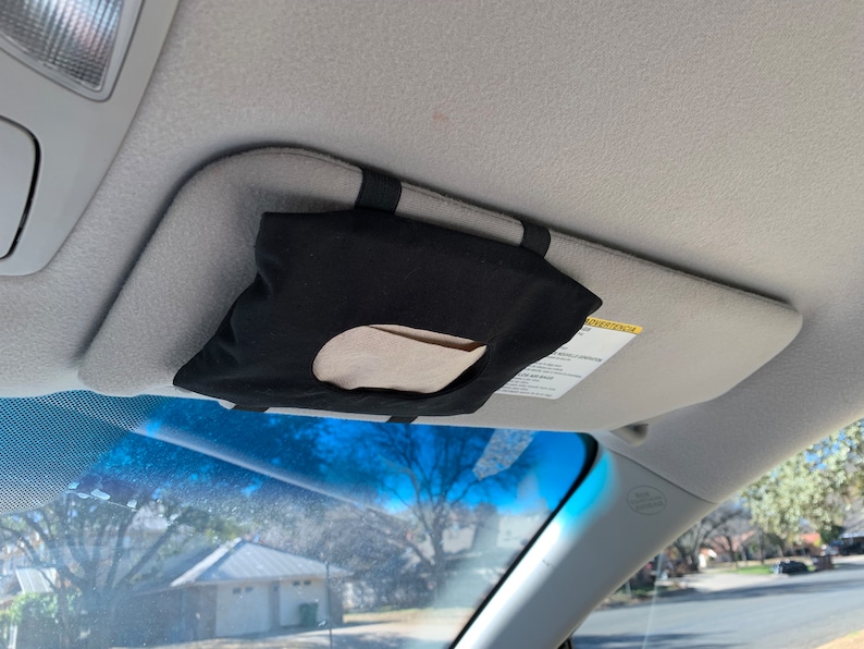 Op de afbeelding: Een zwarte stoffen tissuehouder met een wit tissue erin, bevestigd aan de zonneklep van een auto.