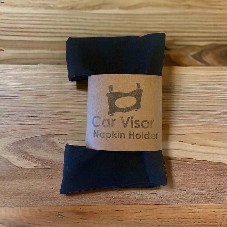 Op de afbeelding: Zwarte autozonneklep-servetthouder met een bruin papieren etiket met de tekst "Car Visor Napkin Holder".