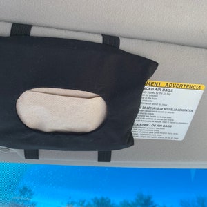 Op de afbeelding: Zwarte stoffen autozonneklep tissuehouder met een wit papiertje erin. De zonneklep heeft een geel waarschuwingslabel met de tekst "Advanced Air Bags" en "ADVERTENCIA" in het Spaans.