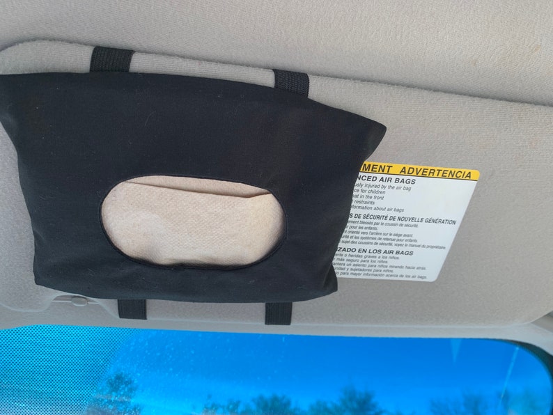Op de afbeelding: Zwarte stoffen autozonneklep tissuehouder met een wit papiertje erin. De zonneklep heeft een geel waarschuwingslabel met de tekst "Advanced Air Bags" en "ADVERTENCIA" in het Spaans.