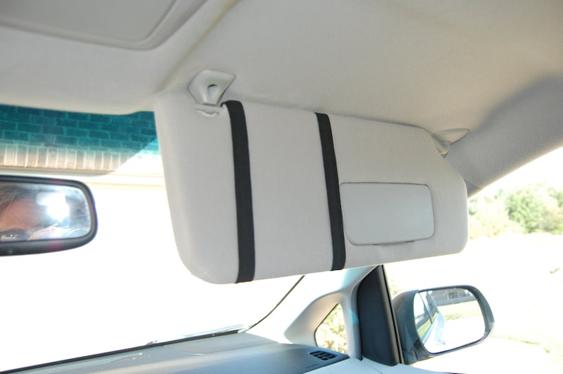 Op de afbeelding: Een witte autozonneklep met twee zwarte banden. De zonneklep is bevestigd aan het plafond van de auto.