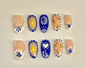 May include: A set of ten press-on nails with various designs. Some nails are nude with white and blue floral accents. Others feature a sun, a white flower, a heart, and orange fruit designs on a blue background. The nails are oval-shaped.