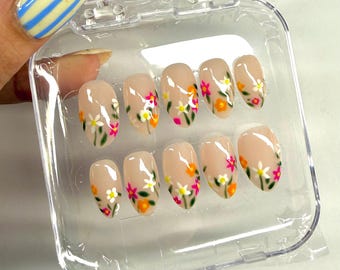 May include: A set of artificial nails in a clear plastic case. The nails are oval-shaped with a nude base and feature a floral design with white, pink, and orange flowers. The nails are held by a person with a painted fingernail.