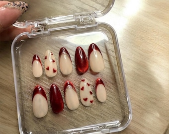 May include: A set of ten false nails in a clear plastic container. The nails are oval shaped and have a red base with a white tip. Some of the nails have a red heart design on the white tip.