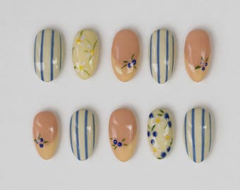May include: A set of ten press-on nails featuring a variety of designs. Some nails have vertical blue stripes on a cream base, while others feature hand-painted floral patterns with white daisies and small blue berries on cream or peach backgrounds.