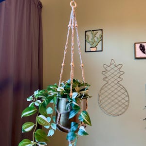 Op de afbeelding: Een macramé plantenhanger met houten kralen, die een gouden pot met een groene, bladerige plant vasthoudt. De hanger hangt aan een plafondhaak.