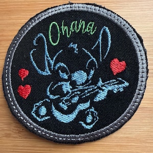 May include: Round black patch with a blue Stitch playing a ukulele, the word "Ohana" in green, and two red hearts. The patch has a gray stitched border and is on a wooden surface.