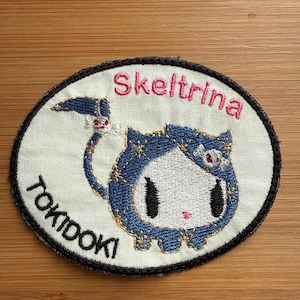 May include: Oval embroidered patch featuring a cartoon cat character with blue and white details, gold accents, and the name "Skeltrina" in pink. The word "Tokidoki" is also embroidered on the patch.
