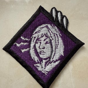 Puede incluir: Parche bordado con un fondo morado en forma de diamante y un borde negro. Presenta una cara blanca estilizada con cabello blanco. Tres formas de garra se extienden desde la parte superior. Un artículo decorativo.