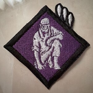 Könnte beinhalten: Ein rautenförmiger Stickaufnäher mit violettem Hintergrund und einer weißen, knienden Figur. Die Figur trägt eine Mütze und strukturierte Kleidung. Ein schwarzer Rand rahmt das Design ein, und drei weiße Krallenspuren ragen aus der oberen rechten Ecke.