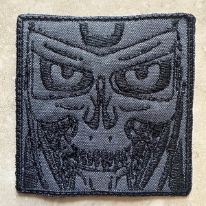 Może przedstawiać: Kwadratowa, szara naszywka z czarnym haftowanym wzorem czaszki. Czaszka ma szczegółowe cechy, w tym oczy, nos i zęby. Naszywka ma czarną obwódkę i prawdopodobnie jest przeznaczona na odzież lub akcesoria.