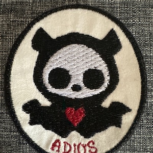 May include: Embroidered patch featuring a black and white cartoon character with a skull-like face, a red heart, and the word "ADIOS" in red. The patch has a white background and a black border.
